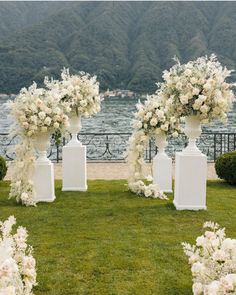 an outdoor ceremony setup with white flowers and tall vases on the grass near water