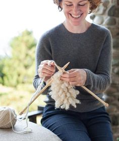 a woman is smiling while knitting yarn on a chair with two wooden skewers