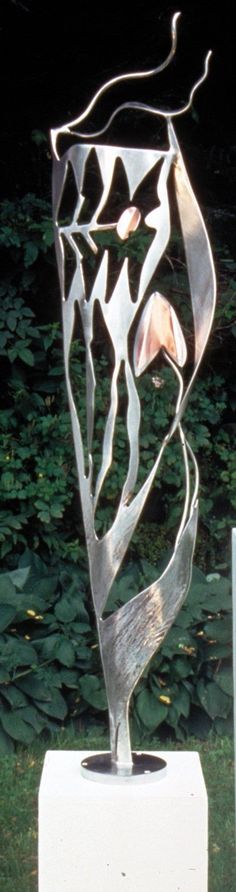 a metal sculpture sitting on top of a white block in front of some grass and bushes