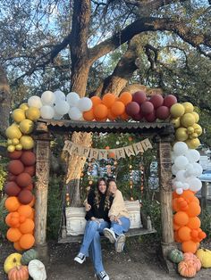 two women sitting on a bench in front of a tree with balloons and pumpkins