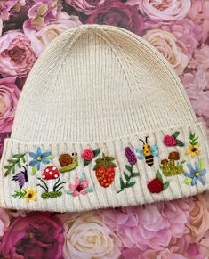 a white knitted hat with embroidered fruit and flowers on it, sitting in front of pink flowers