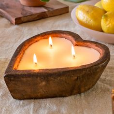 two candles in a heart shaped bowl on a table with lemons and other fruit