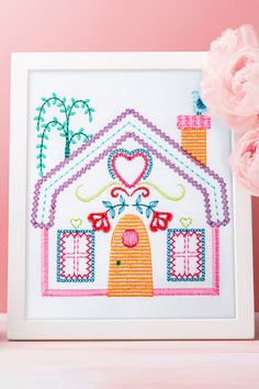 a pink flower sitting next to a white frame with a cross stitch house on it