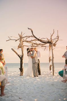 a bride and groom are kissing on the beach under an arch made out of driftwood