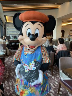 a person dressed as mickey mouse holding a chalk board in front of a dining room