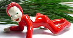 a red figurine laying on its side next to a pine branch with it's eyes closed