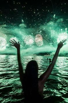 a woman is standing in the water with her arms up as fireworks are set off