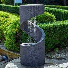 a water fountain in the shape of a spiral on a stone path surrounded by hedges