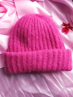 a pink knitted beanie laying on top of a bed next to a flower