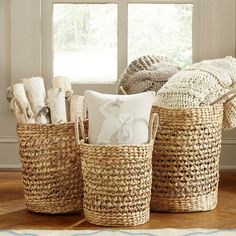 three woven baskets with pillows and blankets in front of a window on a wooden floor