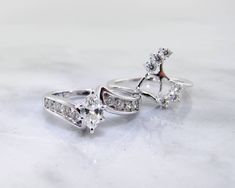 two rings with diamonds on them sitting on a white table top, one is in the shape of an arrow