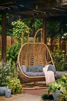 a wicker swing chair with cushions on it in a garden area surrounded by plants