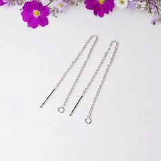 three pairs of silver chains on a white surface with purple flowers in the back ground