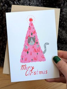 a hand holding up a christmas card with a cat in a tree on it's side