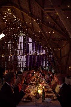 a group of people sitting at a table in a barn with lights on the ceiling