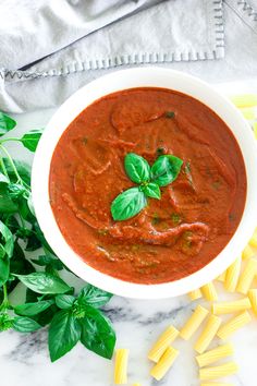 a white bowl filled with red sauce and pasta on top of a marble counter next to green leaves