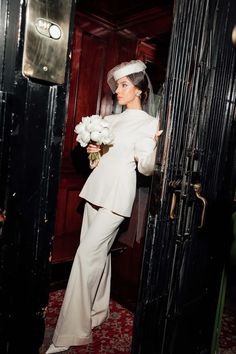 a woman standing in front of a door wearing a wedding dress and holding a bouquet of flowers