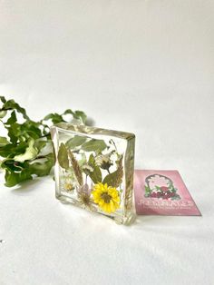 a small glass block with flowers in it and a card next to it on a white surface