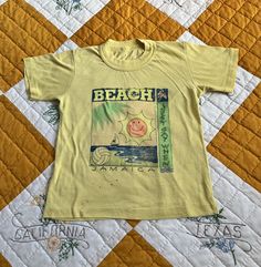 "80s PALE YELLOW CROPPED \"JAMAICA\" GRAPHIC TEE: - Pale yellow tee - Beach scene graphic w/ \"BEACH\" \"JUST SAY WHEN\" + \"JAMAICA\" - Cropped length MEASUREMENTS: Bust: 13 1/2\" (unstretched) (please double)  Sleeve: 5 1/2\" Length: 17\" No size marked THERE IS ABOUT 3\" - 3 1/2\" WORTH OF STRETCH IN THIS WOULD BEST FIT A SIZE EXTRA EXTRA SMALL, EXTRA SMALL OR SMALL  (DEPENDING ON FIT PREFERENCE) CONDITION: Overall, very good conditionthe blue on the graphic is cracking (see 1st photo), there are some black marks on the front-lower-left corner (see 2nd photo) + there is a small pin hole on the back (see 3rd photo).  PLEASE REACH OUT WITH ANY QUESTIONS YOU MAY HAVE. I'M HAPPY TO HELP! ALSO, FEEL FREE TO CHECK OUT MY SHOP'S POLICIES. ITEMS WILL BE CAREFULLY + LOVINGLY PACKAGED + SHIPPED. Cheap Yellow 90s Tops, Green 90s Style T-shirt For Summer, Yellow Crew Neck T-shirt For Beach, Vintage Short Sleeve Tops For Vacation, Retro Yellow Beach Top, Yellow Tops With Vintage Print For Summer, Summer Crew Neck T-shirt With Vintage Print, Summer Vintage Print Crew Neck T-shirt, Retro Yellow Shirt For Vacation