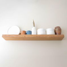 a wooden shelf filled with cups and vases on top of a white wall next to a potted plant