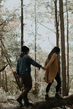 a man and woman holding hands walking across a fallen tree in the middle of a forest