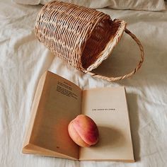 an open book sitting on top of a bed next to a basket and two peaches