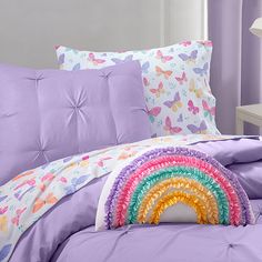 a bed covered in purple sheets and pillows with a rainbow on the pillow case next to it