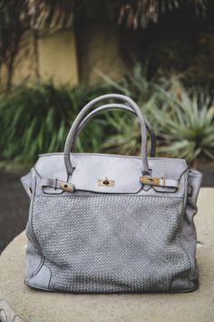 The Rosalind tote in grigio (gray). Front view sitting on tabletop. Vintage, soft woven leather with brass clasp & lock. Luxury Satchel With Braided Handles, Luxury Satchel With Braided Handles For Travel, Luxury Satchel Tote For On-the-go, Luxury Bags With Braided Double Handles, Luxury Shoulder Bag With Braided Double Handles, Luxury Weekender Bag For On-the-go, Luxury Satchel With Rolled Handles For On-the-go, Elegant Travel Bag With Double Handle For Daily Use, Luxury Tote Shoulder Bag With Braided Handles