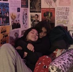 two people sitting on a couch with posters all over the wall behind them and one person hugging her