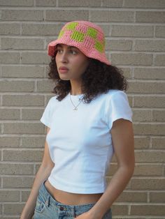 a woman wearing a pink and green crochet hat standing in front of a brick wall