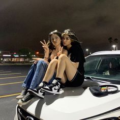 two women sitting on the hood of a white car in front of a parking lot