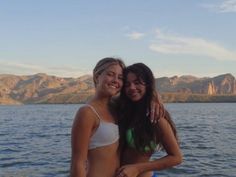 two young women standing next to each other near the water with mountains in the background