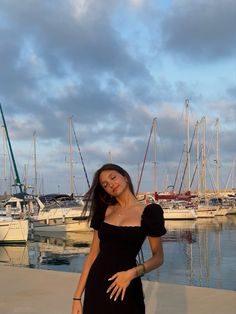 a woman in a black dress posing for the camera near some boats at a marina