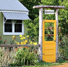 a yellow door in front of a green house with flowers and trees around it that says welcome