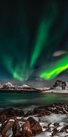 the northern lights shine brightly over snow covered rocks and water in front of snowy mountains