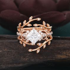 a gold ring with a princess cut diamond surrounded by leaves and diamonds on top, sitting on a piece of wood