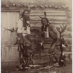 two native american men standing next to each other in front of a log cabin with an open door