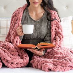 a woman sitting on a bed holding a coffee cup and a book in her hands