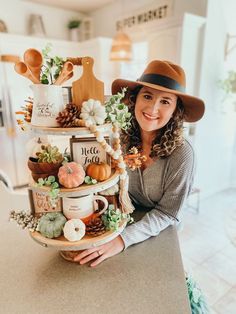 a woman wearing a brown hat holding a tiered tray filled with fall decorations and pumpkins