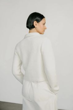 a woman standing in front of a white wall wearing a white sweater and pants with pockets