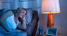 a woman laying in bed talking on her cell phone and smiling at the camera with a lamp next to her