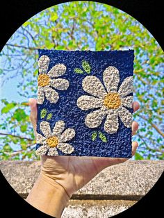 a hand holding up a piece of paper with white flowers on it and green leaves in the background