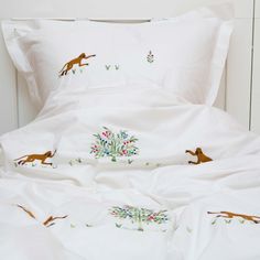 a bed with white sheets and embroidered animals on the pillowcase, along with two pillows