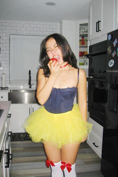 a woman in a yellow tutu skirt eating an apple while standing in a kitchen