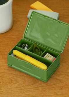 a green box with compartments holding pens and other office supplies on a wooden table next to a cup