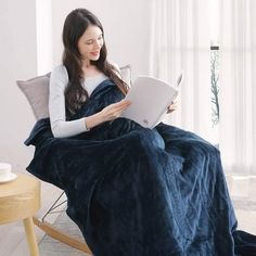 a woman sitting on a chair reading a book while wearing a blue blanket and holding a laptop