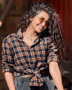 a woman with long curly hair wearing sunglasses and a checkered shirt smiles at the camera