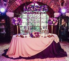 a table with purple flowers and candles on it in front of a brick wall that has an arched window