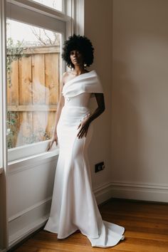 a woman standing in front of a window wearing a white dress with an off the shoulder neckline