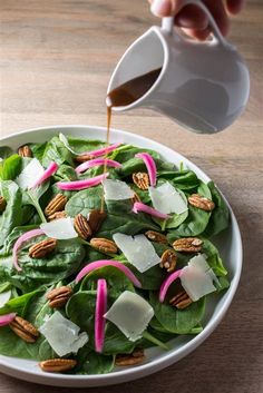 a person pouring dressing onto a salad in a white bowl on a wooden table with spinach leaves and pecans